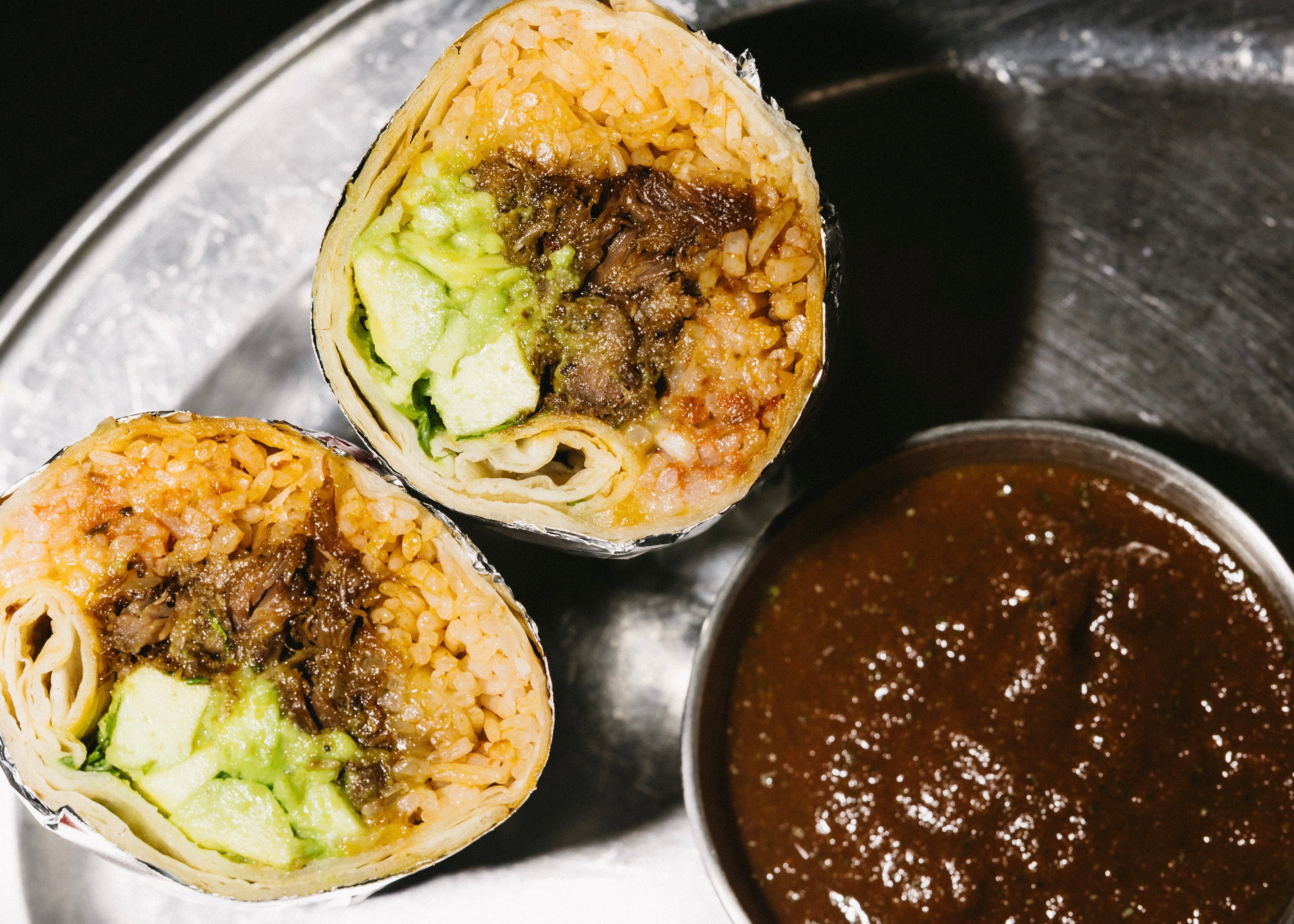 Two burritos cut in half on a metal tray with a side of sauce.